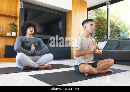 Méditer divers amis masculins assis les jambes croisées dans un salon moderne, avec des tapis de yoga noirs Banque D'Images