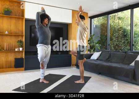 Équilibrer divers amis masculins tenant l'arbre pose dans le salon moderne, avec des tapis de yoga noirs Banque D'Images