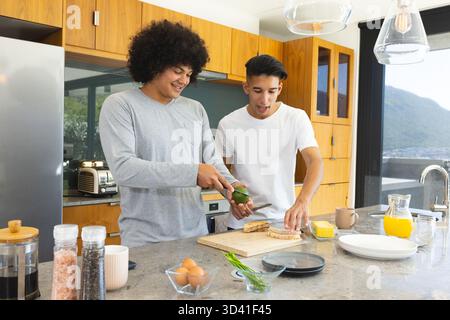 Hacher l'avocat, divers amis masculins préparant le petit déjeuner à l'îlot de cuisine avec planche à découper Banque D'Images