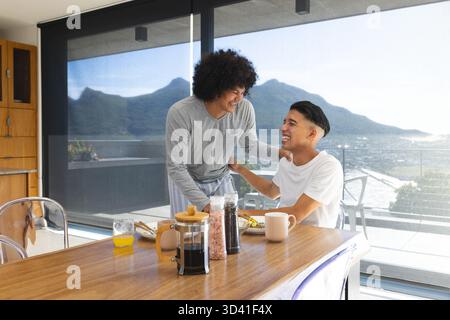 Rire divers partenaires masculins partageant le petit déjeuner à la table à manger à la maison, avec café presse français Banque D'Images