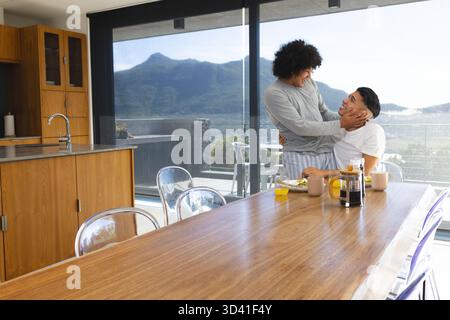Embrasser divers partenaires masculins debout par une table en bois avec du jus d'orange dans la cuisine, espace de copie Banque D'Images