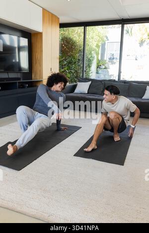 Étirer divers amis masculins tenant des poses de yoga dans un salon moderne, avec des tapis de yoga noirs Banque D'Images