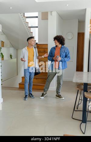 Debout divers amis masculins discutant au pied de l'escalier dans la maison ouverte, tenant smartphone Banque D'Images