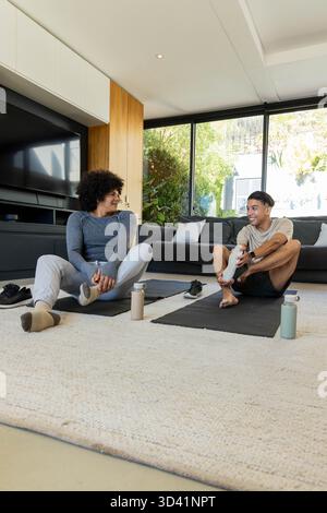 Étirer divers amis masculins tenant les pieds sur des tapis de yoga dans le salon moderne, avec des bouteilles d'eau Banque D'Images
