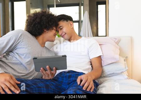 Couple masculin diversifié partageant un moment tendre sur le lit à la maison, tenant la tablette Banque D'Images