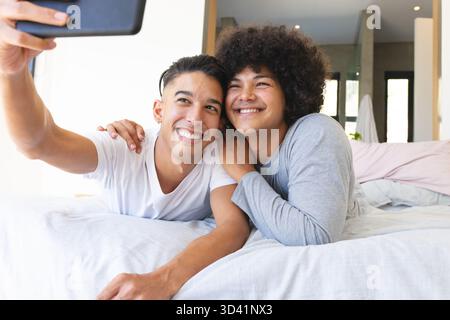Couché divers partenaires masculins câlins sur le lit dans une chambre moderne, tenant la tablette pour selfie Banque D'Images