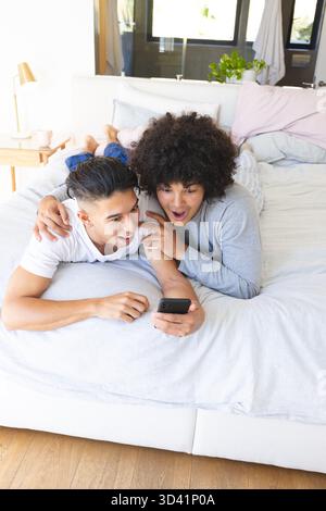 Rire divers amis masculins allongés sur le lit à la maison, avec smartphone, oreillers pastel et lampe Banque D'Images