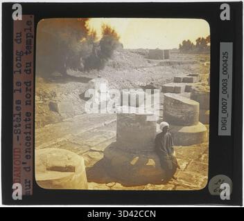 Cette image représente un ouvrier assis à côté de colonnes le long du mur extérieur nord du temple de Montu à Medamud, comme on le voit sur une diapositive en verre de 1839-1939. La scène reflète les détails architecturaux et le cadre de l'ancien temple. Banque D'Images