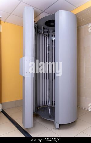 Un lit de bronzage vertical moderne, gris et vide, également connu sous le nom de solarium ou de transat, est montré avec sa porte ouverte dans une salle de salon carrelée aux murs jaunes. Banque D'Images