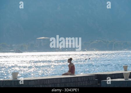 Jeune femme assise sur le front de mer relaxant en écoutant de la musique dans la lumière du soleil du matin dans la baie de Kotor au Monténégro Banque D'Images