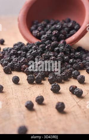vue latérale de grains de poivre noir se déversant d'un pot en terre cuite sur une surface rustique en bois, nature morte terreuse douce et concept d'épices culinaires Banque D'Images