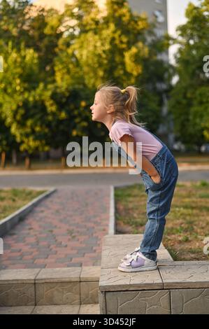 Fille en combinaison en denim et T-shirt rose penchée en avant, regardant vers le bas, debout sur le mur bas. fille regarde vers le bas de trottoir sur fond de parc. vert Banque D'Images