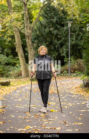 Une femme marche en toute confiance avec des bâtons de marche nordique le long d'un chemin bordé d'arbres dans un parc. Des feuilles d'automne brillantes recouvrent le sol, créant un fond vibrant Banque D'Images