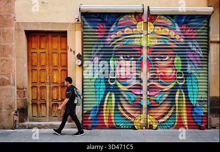 Homme marchant devant une murale de rue vibrante représentant un visage de guerrier indigène peint sur un volet de vitrine en métal dans un cadre urbain Banque D'Images