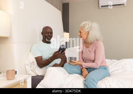 Montrant smartphone couple senior diversifié assis sur le lit dans la chambre, avec tasses horloge numérique Banque D'Images