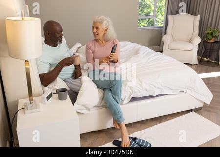 Discuter couple senior diversifié assis sur le lit dans la chambre, avec tasses à café et smartphone Banque D'Images