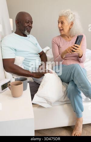 Partage de l'écran de smartphone couple senior divers assis sur le lit dans la chambre, avec tasses à café Banque D'Images