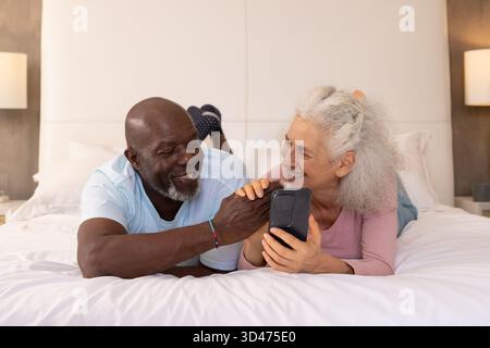 Couché couple senior diversifié sur un lit king-size dans une chambre moderne, partage smartphone Banque D'Images