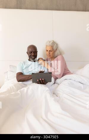 Lecture couple senior diversifié assis dans un lit blanc, tenant tablette et tasse en céramique Banque D'Images