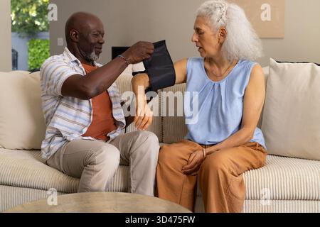 Couple senior diversifié adaptant le brassard de pression artérielle sur le bras dans le salon, avec canapé beige Banque D'Images
