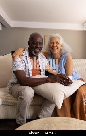 Couple senior souriant assis près du canapé dans le salon, avec table basse et verres clairs Banque D'Images