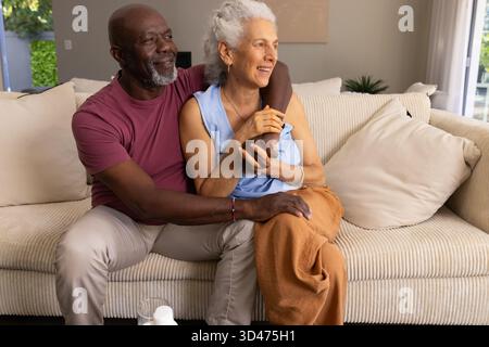 Sourire couple senior diversifié assis étroitement sur le canapé beige dans le salon, avec table basse Banque D'Images
