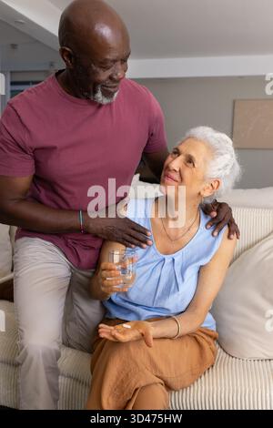 Couple senior diversifié partageant moment dans le salon, avec canapé, pilule et verre d'eau Banque D'Images