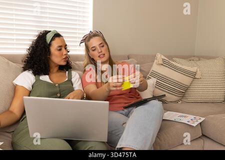 Collaborer diverses amies féminines assises sur un canapé beige dans le salon, avec ordinateur portable et graphiques Banque D'Images