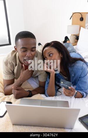 Parcourir le couple afro-américain shopping en ligne sur le lit dans une chambre lumineuse, avec ordinateur portable et carte de crédit Banque D'Images