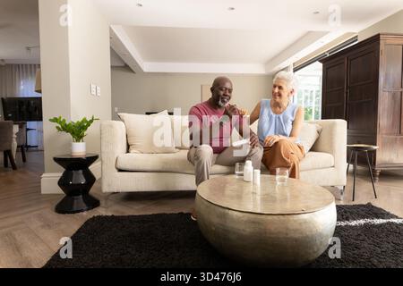 Couple senior diversifié prenant des médicaments sur le canapé à la maison, avec des bouteilles de pilules blanches et des verres à eau Banque D'Images