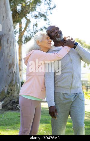Couple senior diversifié s'embrassant sous un grand arbre dans un parc urbain, avec smartwatch noire Banque D'Images