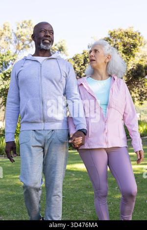 Marcher un couple diversifié tenant la main à travers l'herbe du parc ensoleillée, avec smartwatch et vêtements de sport Banque D'Images