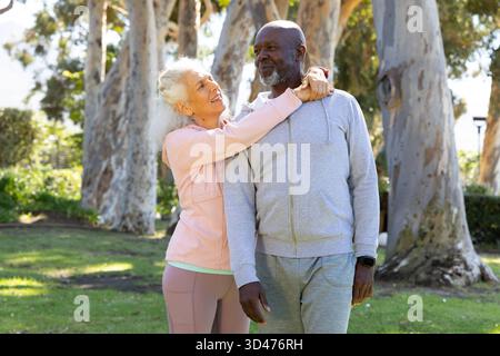 Embrassant un couple diversifié debout sur l'herbe verte dans le parc ensoleillé, avec smartwatch visible Banque D'Images