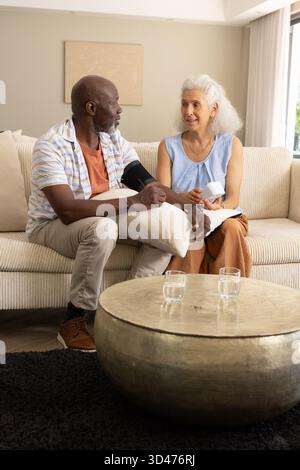 Couple senior ayant la lecture de la pression artérielle dans le salon, avec brassard, moniteur et verres à eau Banque D'Images