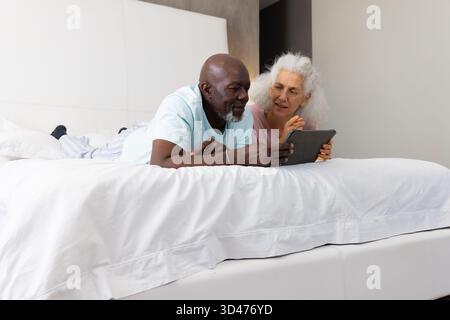 Couché couple senior diversifié sur un lit king-size dans une chambre moderne, à l'aide d'un ordinateur tablette Banque D'Images