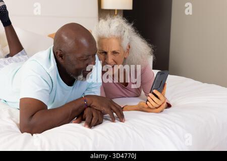 Relaxant couple senior diversifié s'inclinant sur le lit dans la chambre, avec smartphone, oreillers et lampe Banque D'Images
