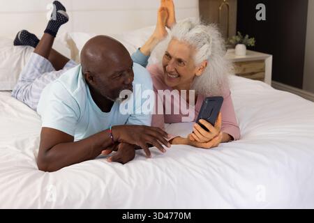 Pointant un couple senior diversifié à l'aide d'un smartphone dans une chambre confortable, avec une lampe moderne et une plante en pot Banque D'Images