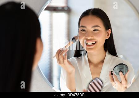 Joyeuse jeune femme japonaise applique Blush tout en souriant dans le miroir de salle de bain Banque D'Images