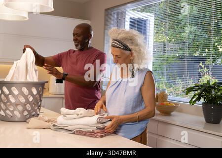 Couple senior diversifié pliant et triant le linge propre sur l'îlot de cuisine, avec panier à linge gris Banque D'Images