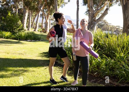 Marchant diverses amies de sexe féminin discutant tout en se promenant dans le parc ensoleillé, avec des tapis de yoga et des bouteilles Banque D'Images