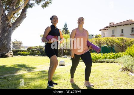 Promenez diverses amies transportant des tapis de yoga sur la pelouse ensoleillée, avec une maison au toit carrelé derrière Banque D'Images