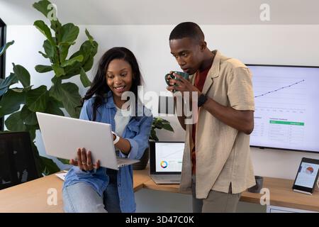 Collègues afro-américains se penchant sur un ordinateur portable au bureau, avec des moniteurs affichant des graphiques Banque D'Images