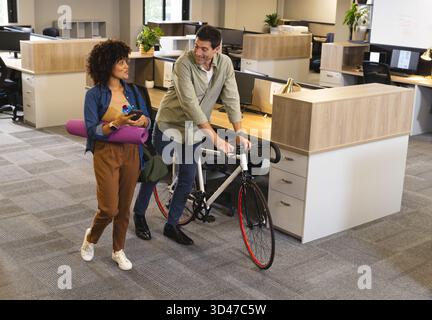 Marcher divers collègues transportant du matériel de fitness à travers le bureau moderne, avec vélo et tapis de yoga Banque D'Images