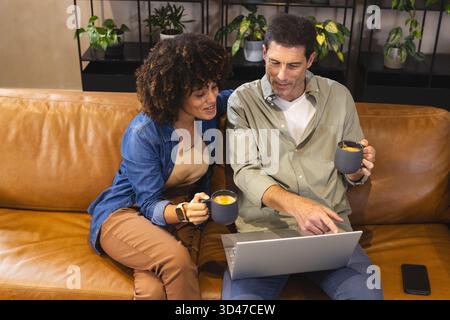Assis couple divers tenant des tasses regardant l'écran d'ordinateur portable sur le canapé en cuir à la maison, avec des plantes Banque D'Images