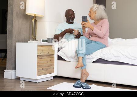 Siroter du café couple senior diversifié assis sur le lit à la maison, tenant smartphone et tasse à café Banque D'Images