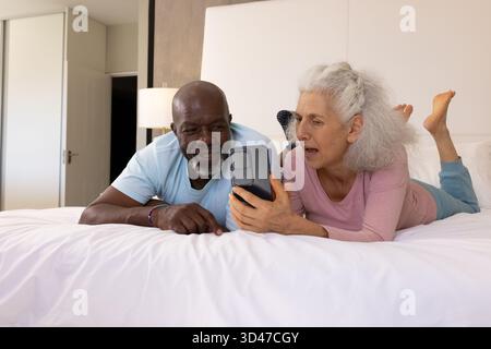 Rire couple senior diversifié couché sur le lit avec des draps blancs dans la chambre, avec smartphone Banque D'Images
