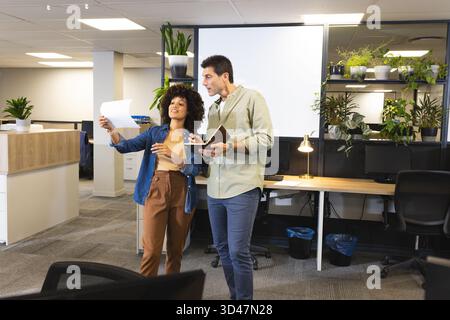 Discuter de divers collègues examinant des documents imprimés dans un bureau ouvert, avec des écrans d'ordinateur Banque D'Images