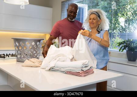 Divers couples seniors pliant des draps ensemble dans une cuisine moderne, avec panier à linge et plantes Banque D'Images