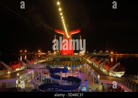 Vue nocturne sur le pont d'un bateau de croisière avec toboggan éclairé, entonnoir rouge lumineux, canots de sauvetage alignés et lumières reflétant sur l'eau calme du port Banque D'Images