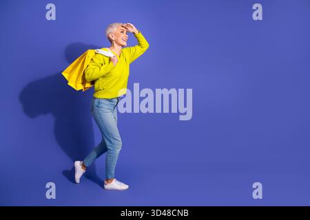 Femme mature énergique avec les cheveux blonds courts portant des sacs à provisions jaunes se tient dans un fond de studio violet vif souriant et regardant vers l'avenir Banque D'Images